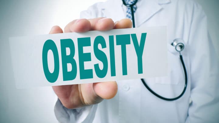A doctor in a white coat holds a sign with the word "OBESITY" in bold green letters, highlighting the long history of obesity awareness and its impact on public health.
