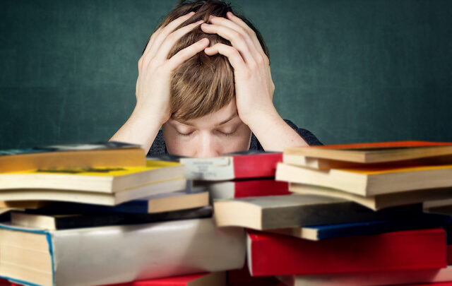 A person sits with their head in their hands behind a stack of books, the weight of test anxiety evident against a dark background.