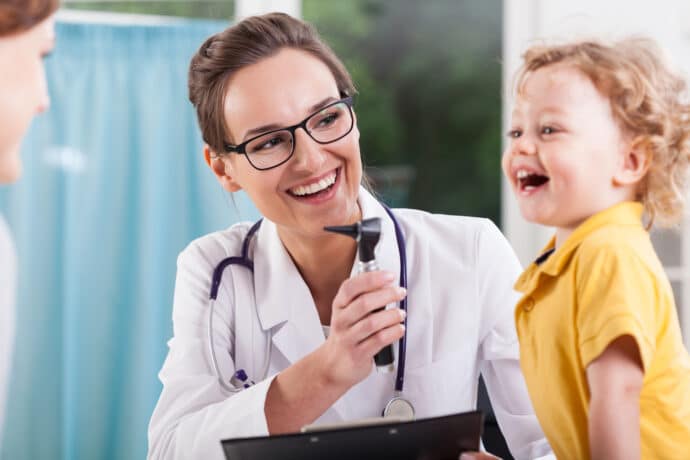 A doctor with glasses smiles while holding a medical instrument, facing a happy child in a yellow shirt.