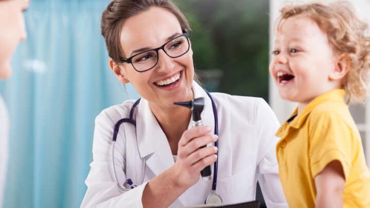 A doctor with glasses smiles while holding a medical instrument, facing a happy child in a yellow shirt.