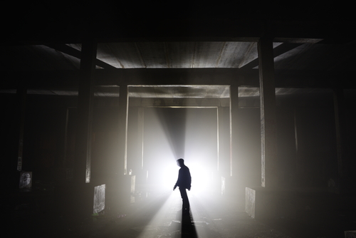 In the silhouette of a person standing in an abandoned, dimly lit concrete structure, it seems as if they are caught in a haunting moment that evokes Cotard's delusion, with beams of light cutting through the darkness like fragments of reality.