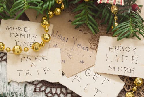 Handwritten New Year's resolutions on paper: "More family time," "Take a trip," "Get fit," and "Enjoy life more," surrounded by pine branches and golden decorations.
