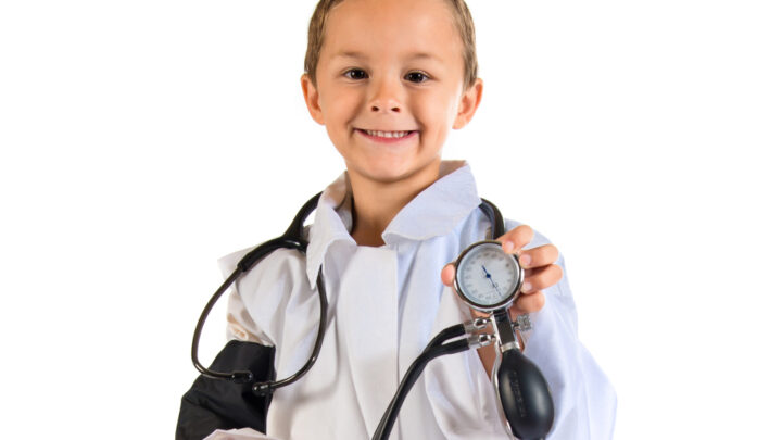 A child in a doctor's coat with a stethoscope holds a sphygmomanometer, smiling at the camera. The scene evokes childhood memories of dreaming to heal, set against a crisp white background.