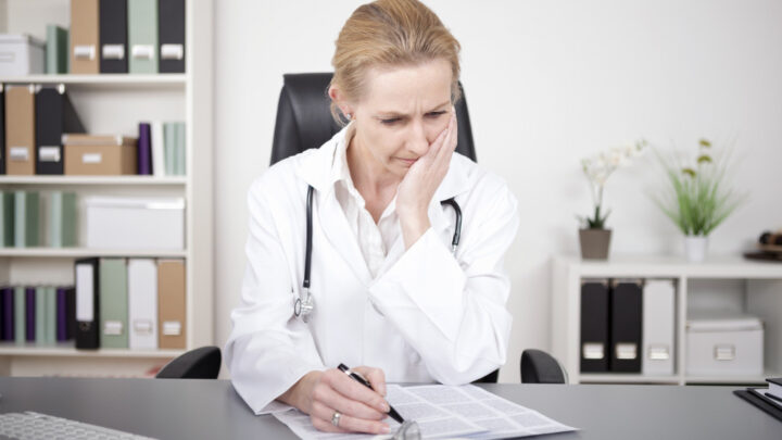 A US physician in a white coat sits at a desk, looking concerned while reviewing documents related to the opioid crisis. Shelves with files and plants are in the background.