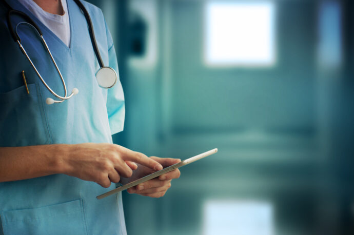 A healthcare professional in scrubs uses a tablet with a stethoscope around their neck in a clinical setting, tapping into the power of medical crowdsourcing. Through platforms like Sermo, solutions are solved collaboratively by connecting with experts worldwide.