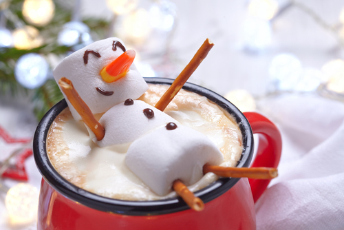 A festive marshmallow snowman with pretzel limbs and a candy nose sits in a red mug of steaming hot chocolate, adding a touch of holiday cheer.