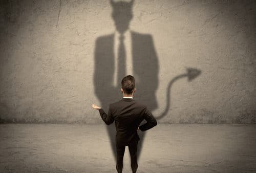 A man in a suit stands in front of a wall, casting an ominous shadow that resembles an evil devil with horns and a tail.