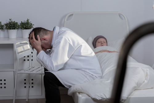 A healthcare professional sits on a chair beside a hospital bed, holding their head in their hands. The weight of imminent death hangs heavy in the room, as the person lies under a blanket, with small potted plants offering a glimpse of life in the background.