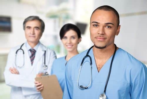 Three healthcare professionals in scrubs and lab coats stand in a hospital, dispelling misconceptions about their roles. The focus is on a man in blue scrubs with a stethoscope, emphasizing his dedication. Two others are in the background, one attentively holding a clipboard.