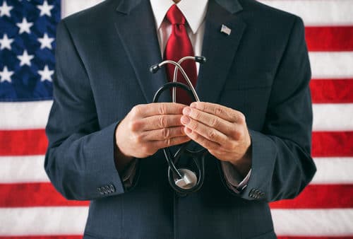 A person in a suit holds a stethoscope, symbolizing adherence to the Goldwater Rule, in front of an American flag.