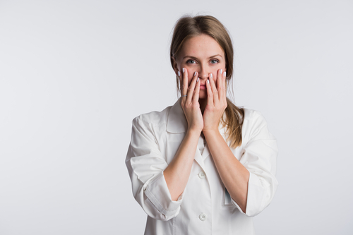 A person in a white lab coat stands with their hands faintly touching their cheeks, gazing forward against a plain background.