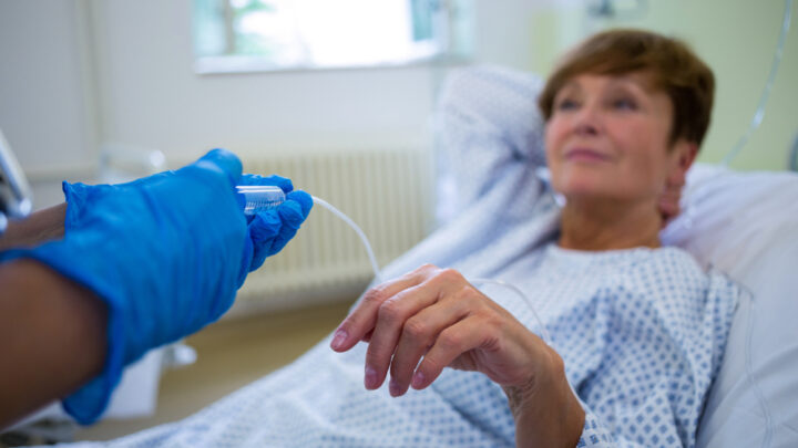 A patient in a hospital bed receiving IV treatment, with someone in blue gloves adjusting the drip as they monitor symptoms.