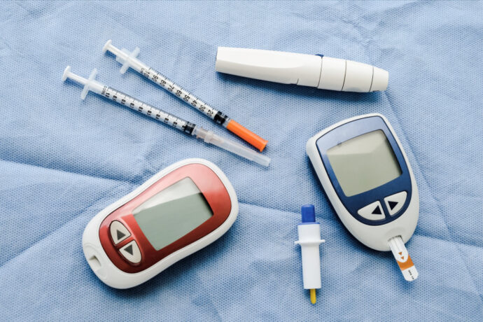 Diabetes management tools on a blue fabric: two blood glucose meters, two syringes with needles, a lancet, and a white lancing device.