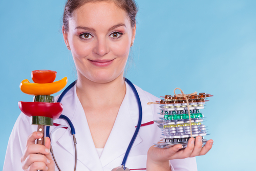 A doctor holds a stack of pills in one hand and a skewer of vegetables in the other, highlighting the choice between diet vs. drugs, while wearing a stethoscope against a blue background.