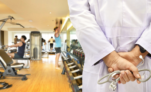 A person in a white lab coat, holding a stethoscope, stands surrounded by exercise equipment in a gym buzzing with people actively working out.