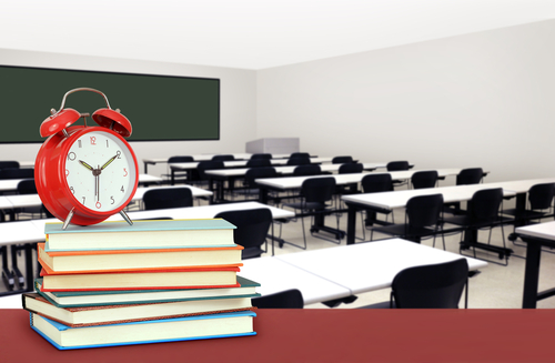 An empty school classroom with rows of desks and chairs, a chalkboard at the front, and in the foreground, a stack of books accompanied by a red alarm clock atop a desk.