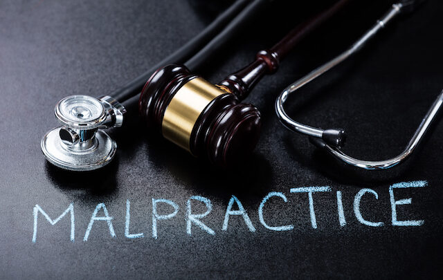 Stethoscope and gavel on a blackboard with the word "Malpractice" written in chalk, hinting at the risk of being sued.