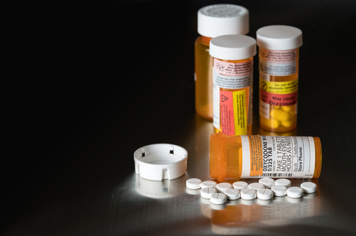 Prescription bottles and pills are scattered on a reflective surface, with one medication bottle lying on its side, spilling several white pills.
