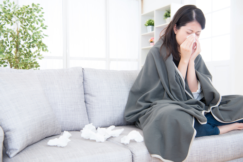 A woman sits on a couch wrapped in a blanket, battling a cold. Using a tissue to blow her nose, she is surrounded by crumpled remains scattered around as evidence of her struggle.