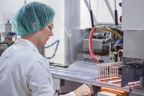 A worker in a lab coat and hairnet operates a machine, enhanced by artificial intelligence, efficiently filling capsules on an assembly line.