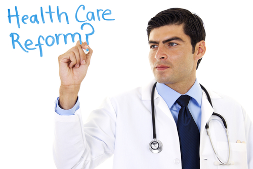 A doctor in a white coat writes "Healthcare Reform?" in blue on a transparent board.