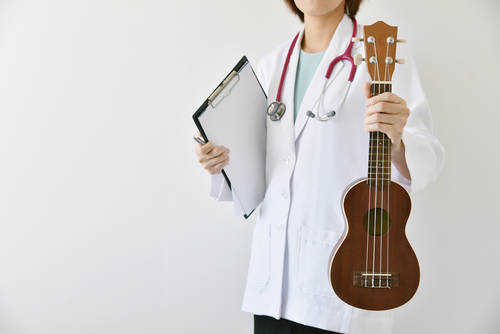 A person in a white coat, resembling an offbeat musician, holds a clipboard and a ukulele.