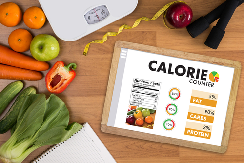 Image of a tablet displaying a calorie counter app, surrounded by a scale, measuring tape, fresh vegetables, vibrant fruits, and a notebook on a wooden surface. The perfect setting for anyone looking to track calories with precision.
