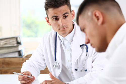 Two doctors in white lab coats are seated at a desk, deep in discussion. One offers professional doctor advice while holding a tablet, the other is focused, glancing down. A stethoscope hangs around one's neck, symbolizing their dedication to medical excellence.