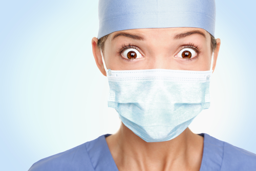 A person in blue scrubs and a surgical mask, possibly working with medical devices, looks directly at the camera with wide eyes against a light blue background.