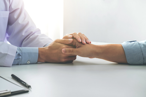 Two people are sitting at a table, where a compassionate doctor in a white coat holds the other person's hand reassuringly. A pen and clipboard are visible on the table.