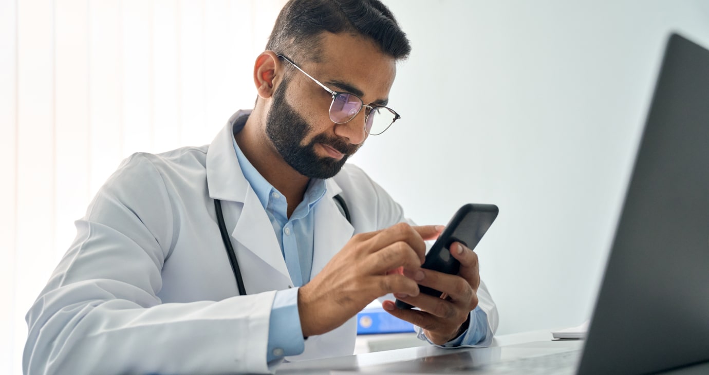Man in a white coat and glasses scrolling through TikTok on his smartphone at a desk, next to an open laptop.