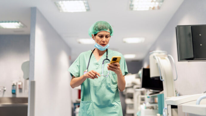 A healthcare professional in scrubs and a hairnet checks their smartphone in a hospital corridor, possibly engaging in some quick social listening amidst the bustle.