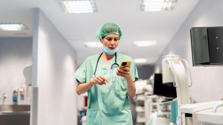 A healthcare professional in scrubs and a hairnet checks their smartphone in a hospital corridor, possibly engaging in some quick social listening amidst the bustle.