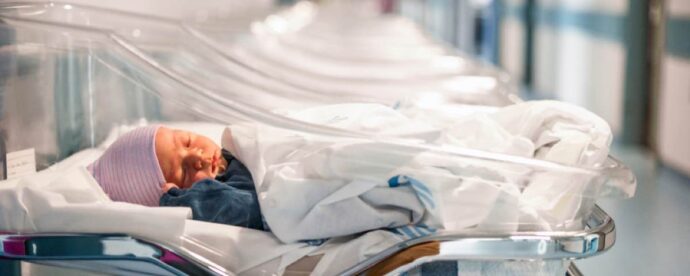 In the July 2019 edition, a newborn baby sleeps peacefully in a hospital crib, wrapped snugly in a white blanket and wearing a striped cap, while a blue outfit peeks through. Several empty cribs are lined up in the background.