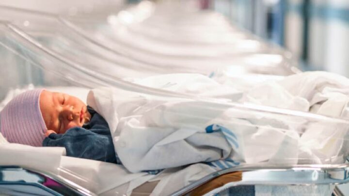 In the July 2019 edition, a newborn baby sleeps peacefully in a hospital crib, wrapped snugly in a white blanket and wearing a striped cap, while a blue outfit peeks through. Several empty cribs are lined up in the background.