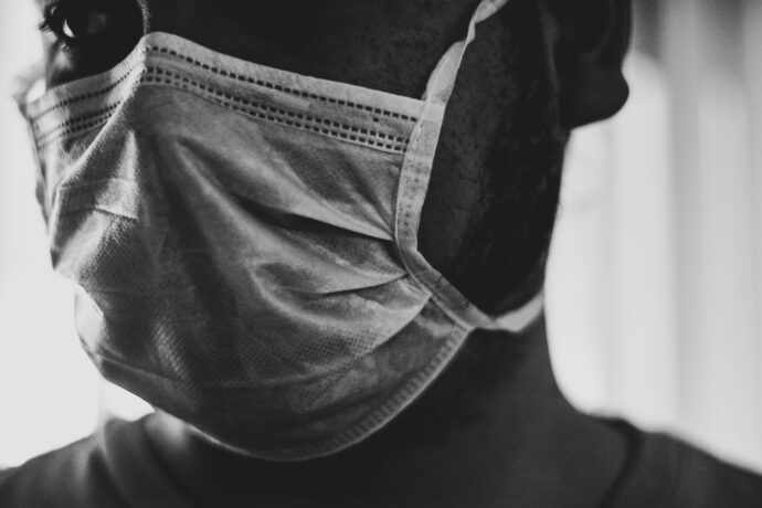 A close-up in black and white highlights a person wearing a face mask, revealing only part of their face and beard, subtly echoing the disparities often seen in hospital care.