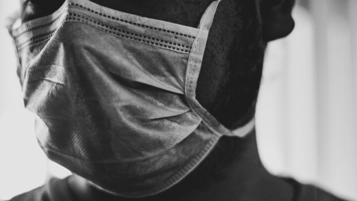 A close-up in black and white highlights a person wearing a face mask, revealing only part of their face and beard, subtly echoing the disparities often seen in hospital care.