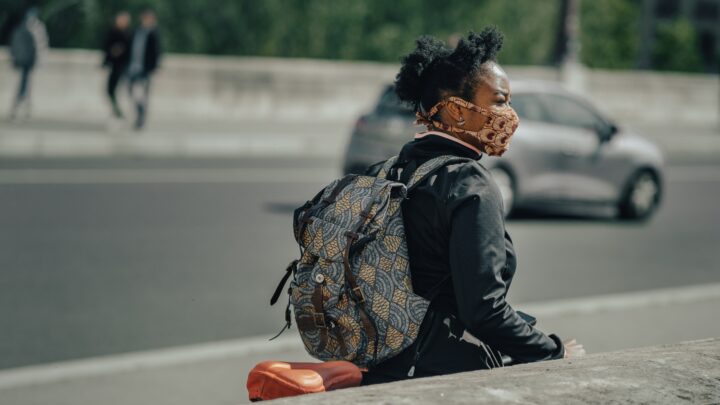 A person wearing a patterned mask and backpack sits on a ledge beside a bustling city street, conscious of the ongoing battle against COVID. Cars zip by, each carrying passengers mindful of their health and immunity in the urban landscape.