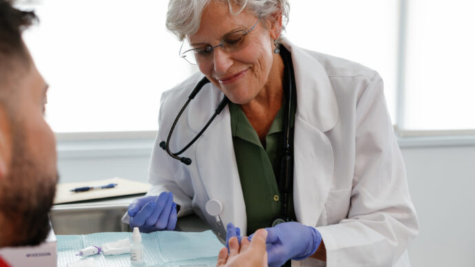 A doctor in a white coat and purple gloves conducts a finger-prick test on a patient, utilizing a small lancet device to ensure accurate results in line with CDC HIV guidelines.