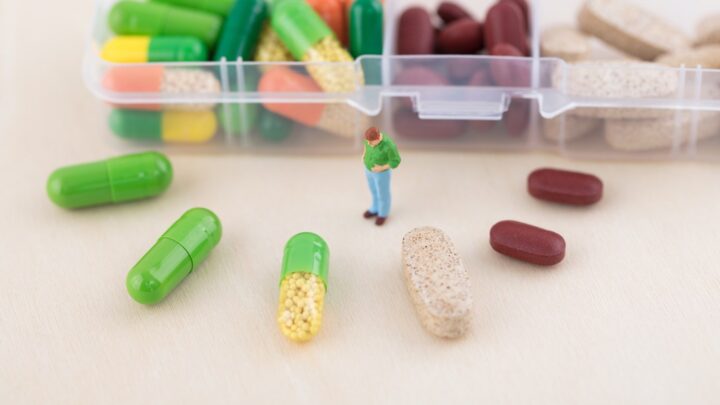 A small figure stands on a table surrounded by large capsules and pills, with an open pill organizer in the background, resembling the vital routine of diabetes patients managing their drug supply.