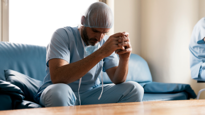 A healthcare worker in scrubs and a hairnet sits on a couch, leaning forward with clasped hands, embodying the burnout problem many physicians face, appearing thoughtful or stressed.