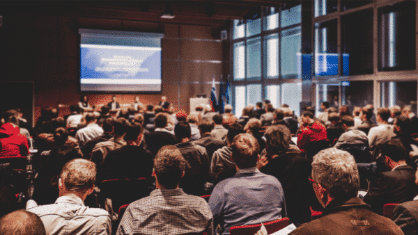 A large audience attentively listens to a panel discussion during an in-person conference on healthcare events, with a projected presentation screen ready.