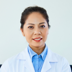 A person with dark hair tied back is wearing a white lab coat and a blue shirt, smiling in front of a plain background, embodying the spirit of health care ethics.