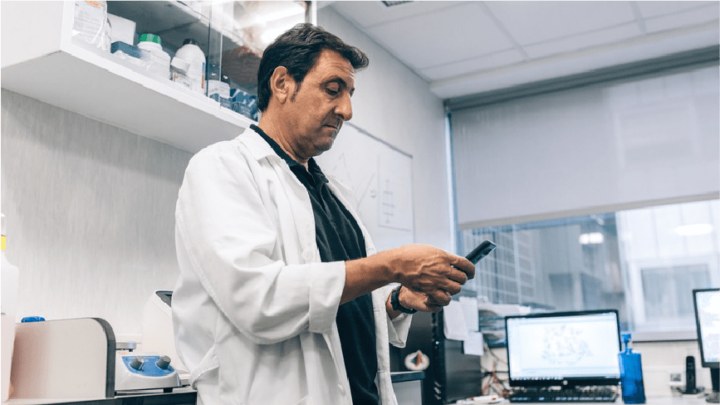 A man in a lab coat stands in a laboratory, looking at a smartphone. Scientific equipment and a computer are visible in the background.