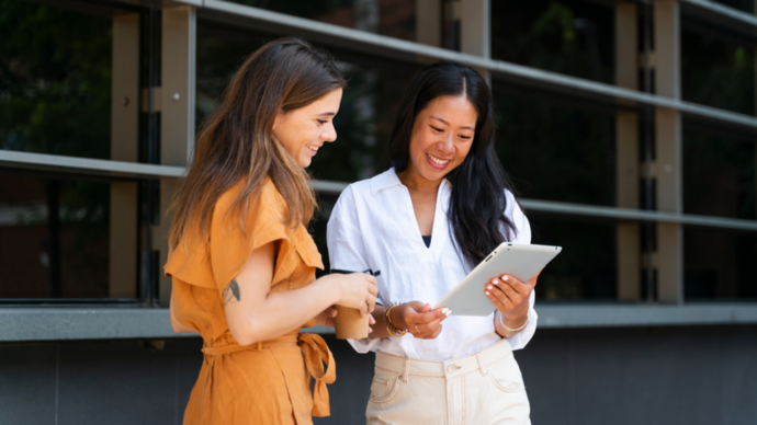 Two women, health system leaders, stand outdoors, smiling as they look at a tablet. One wears an orange dress and holds a coffee cup, while the other is in a white shirt and beige pants. They're in front of a glass building, discussing innovations in US hospital healthcare.