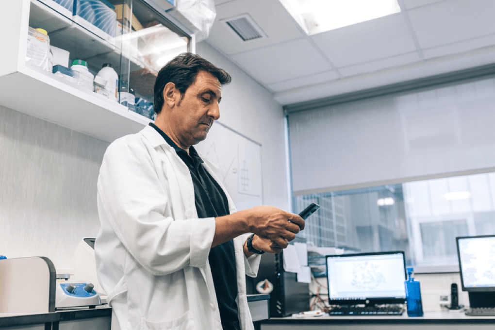 A person in a lab coat checks a device in a laboratory, with computers and lab equipment visible in the background.