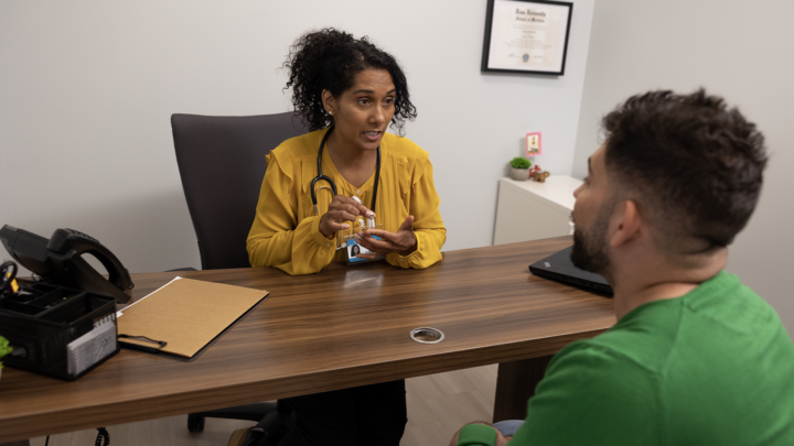 A healthcare professional in a yellow blouse sits at a desk, explaining medication to a seated patient, highlighting its importance in tackling the HIV epidemic. A stethoscope hangs around her neck, and a diploma graces the wall.