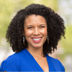 Jessica Knox, wearing a blue shirt and earrings, stands outdoors with a radiant smile. Her curly hair catching the light hints at her vibrant personality.