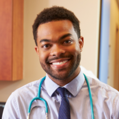 Man with a beard wearing a light blue shirt, tie, and teal stethoscope, smiling in a medical office, embodying the principles of health care law and ethics.