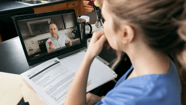 In a telehealth session, a person in blue scrubs engages in a video call with a professional displaying a medication bottle. Open documents are visible on the table, reflecting an exchange of valuable HCP insights.